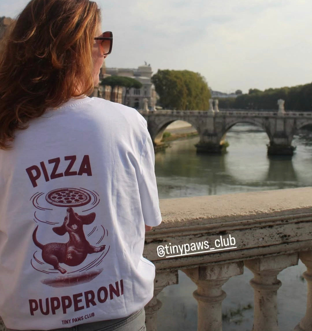 Person wearing a t-shirt with a pizza design and text, standing by a river with a bridge in the background.