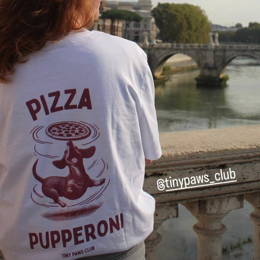Person wearing a t-shirt with a pizza design and text, standing by a river with a bridge in the background.